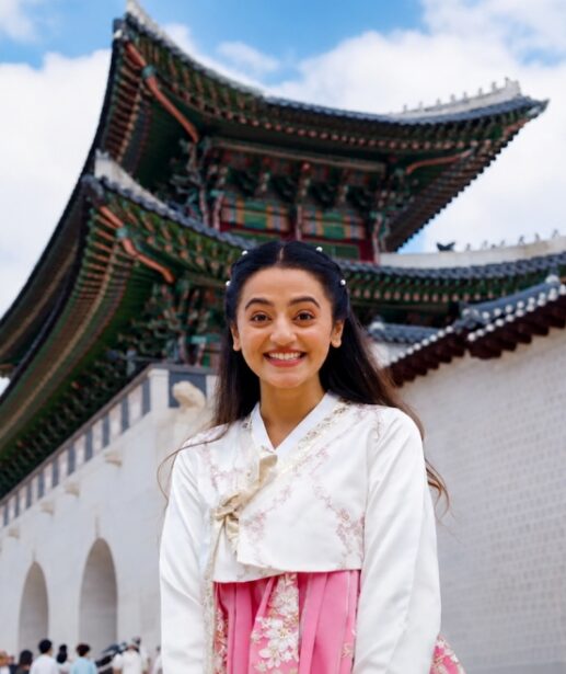 Helly Shah wearing a traditional Korean hanbok at Gyeongbokgung Palace in Seoul, South Korea, smiling in front of the historic palace entrance.