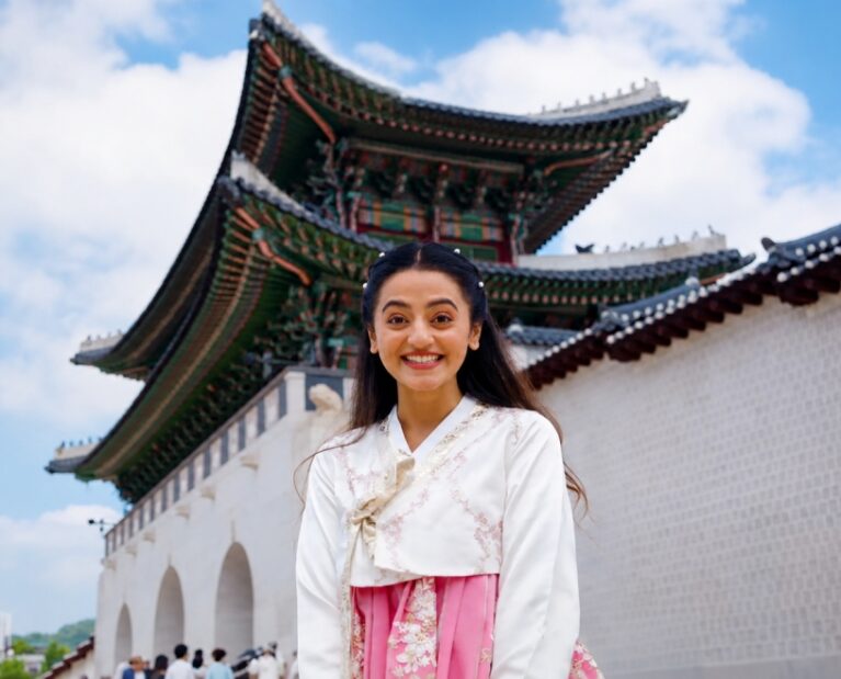 Helly Shah wearing a traditional Korean hanbok at Gyeongbokgung Palace in Seoul, South Korea, smiling in front of the historic palace entrance.