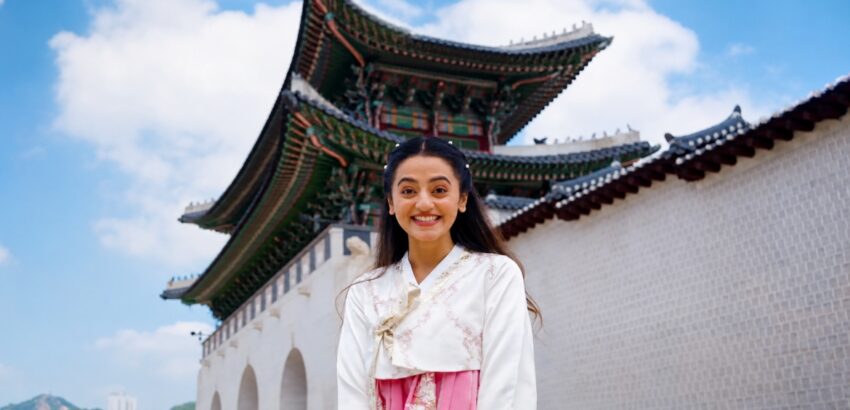 Helly Shah wearing a traditional Korean hanbok at Gyeongbokgung Palace in Seoul, South Korea, smiling in front of the historic palace entrance.