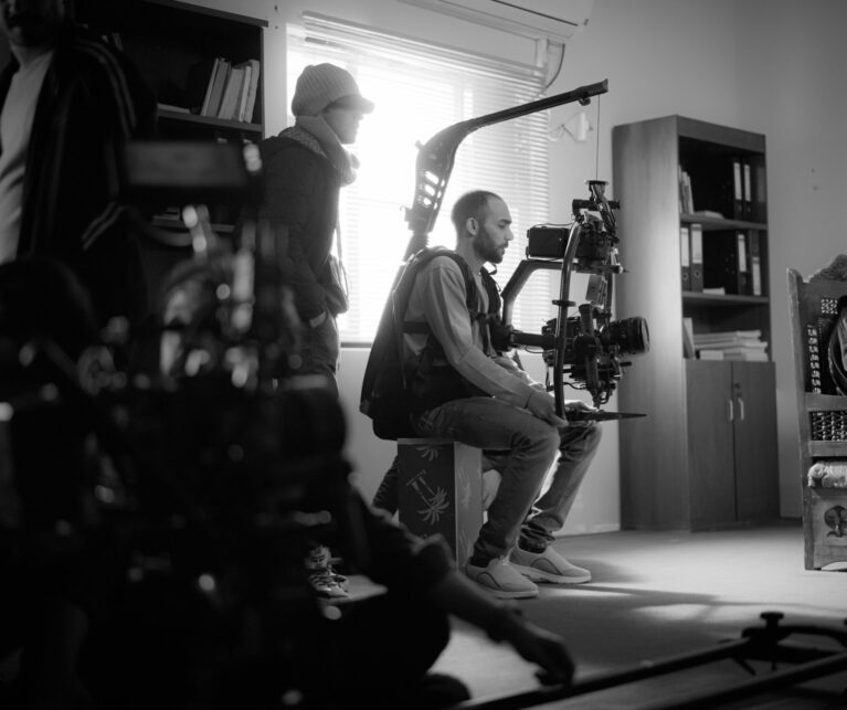 Black and white behind-the-scenes photo of a cinematographer operating a professional camera gimbal rig, filming an elderly bearded man in traditional turban and robes seated in an ornate wooden chair, with soft window light and bookshelf background – illustrating cinematography in action